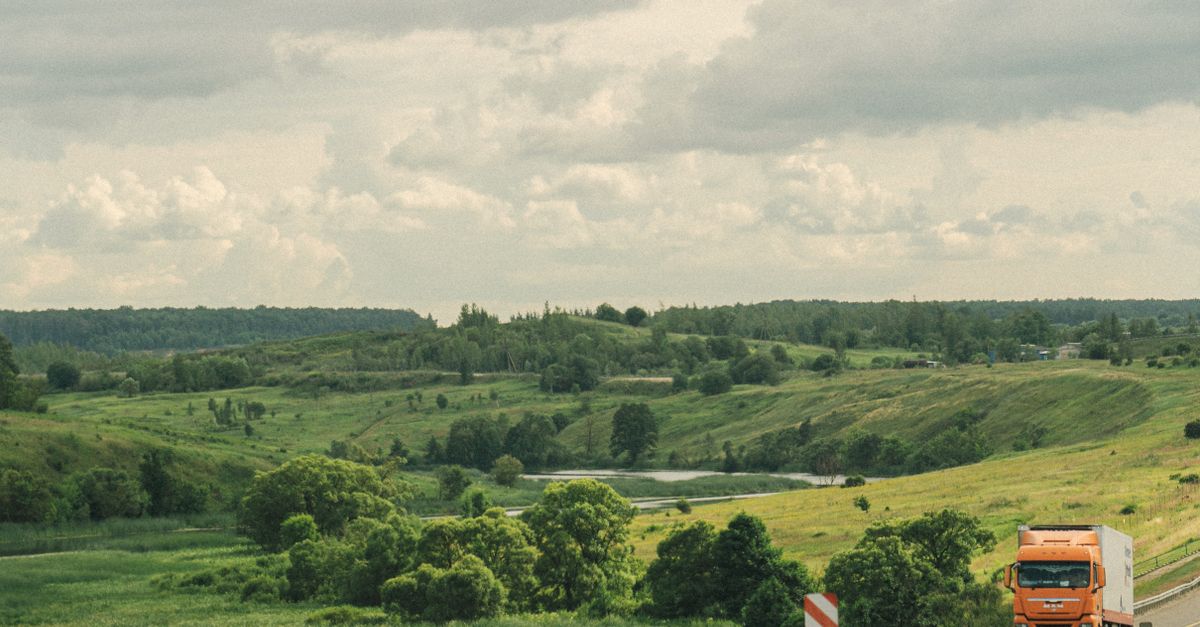 An orange truck drives along a highway through a lush, green, hilly countryside under a cloudy sky.
